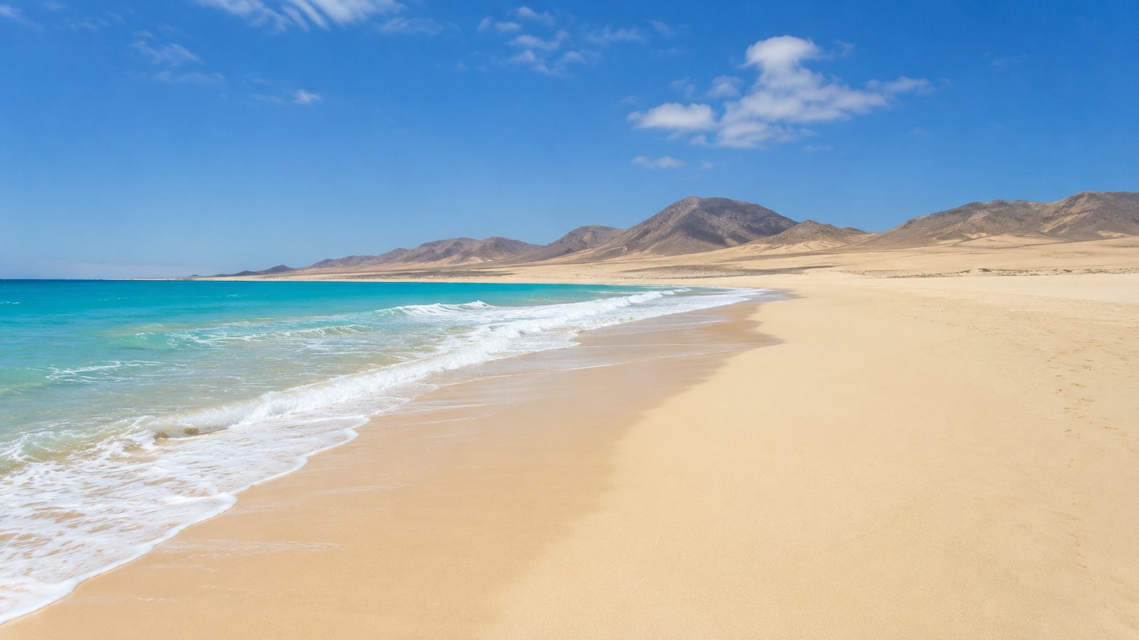 The long, empty white sand of a Boa Vista beach meeting turquoise water below desert mountains, Cape Verde