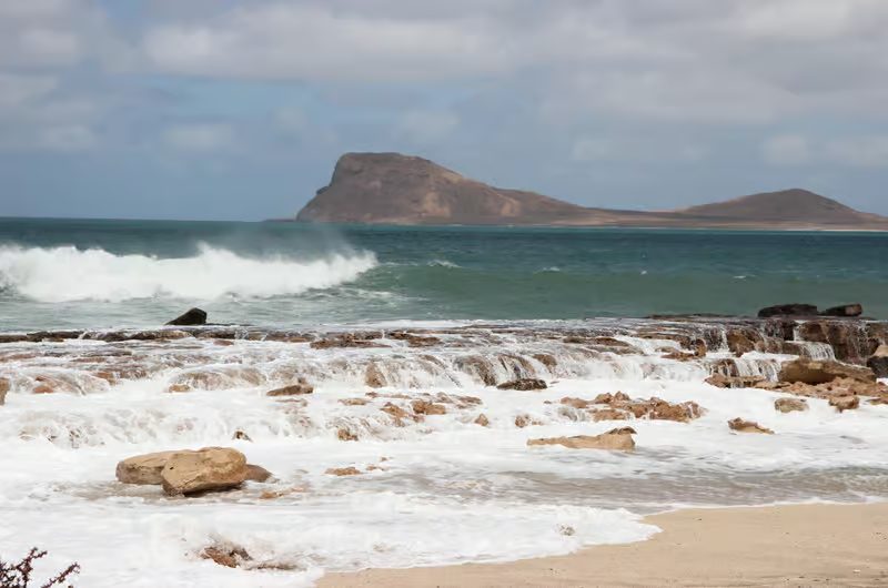 Surf washing over flat rock shelves at the edge of a bay on Sal, Cape Verde, with a brown headland across the water