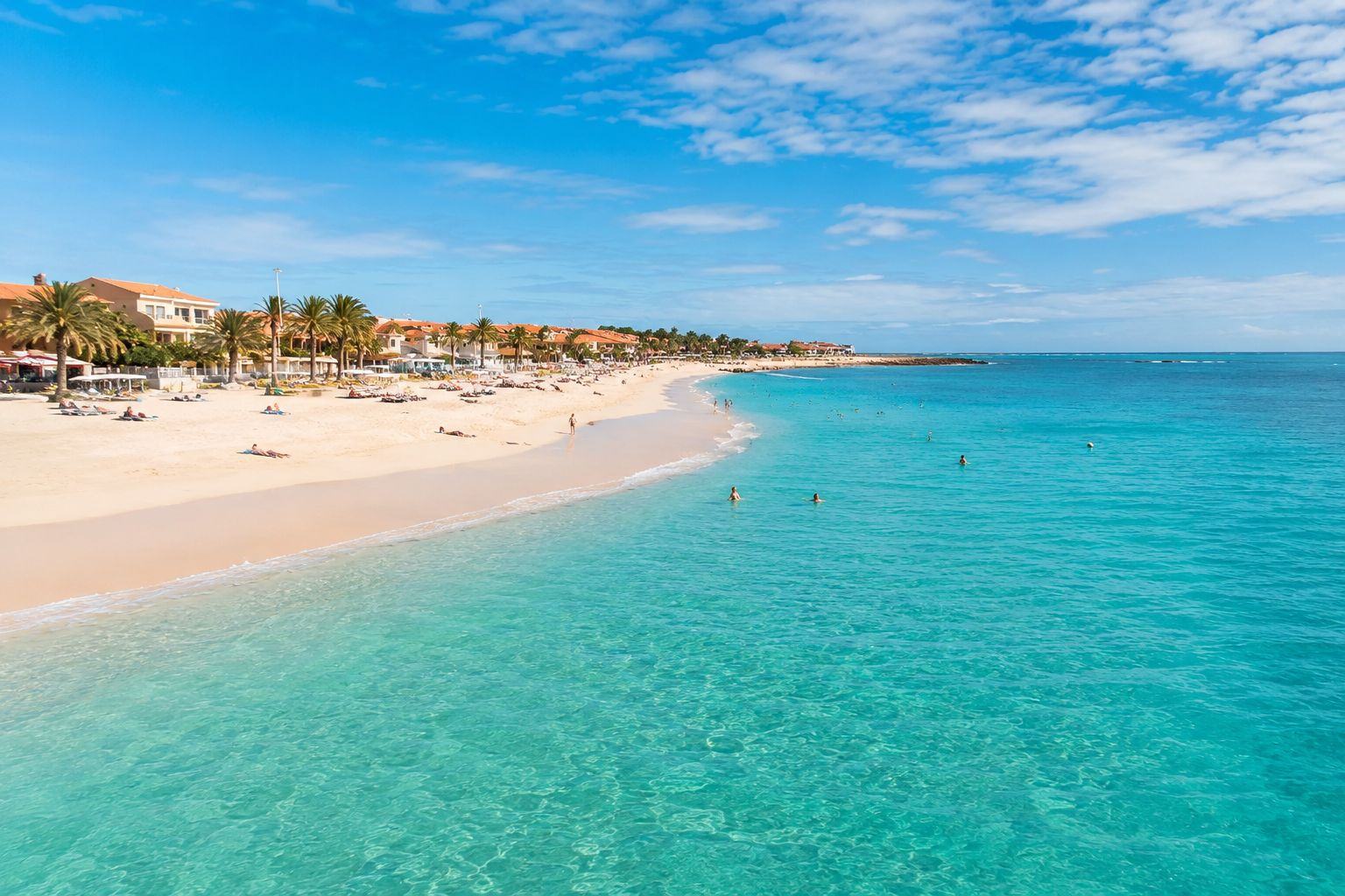 Swimmers in shallow turquoise water at Praia de Santa Maria on Sal, Cape Verde, with sunloungers, palms and low resort buildings along the sand