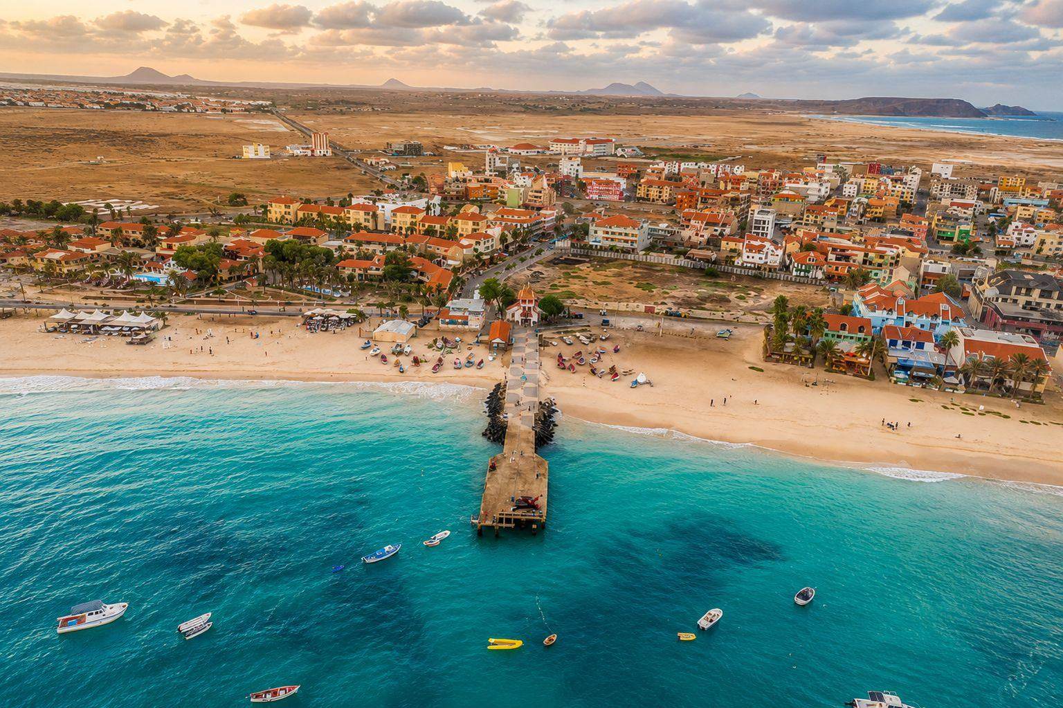 Aerial view of Santa Maria on Sal at sunset, showing the pier reaching into turquoise water, moored boats, the beach and the orange rooftops of the town behind