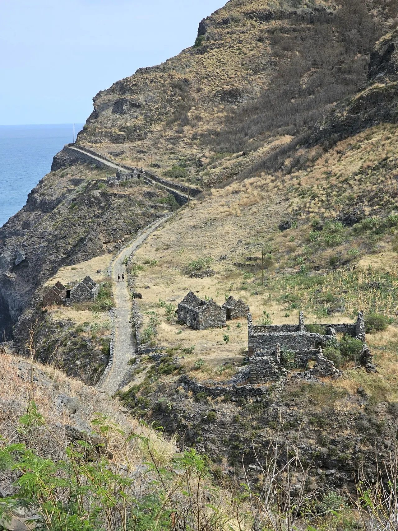 Cobbled coastal path winding past abandoned stone houses on a grassy slope above the sea, Santo Antão