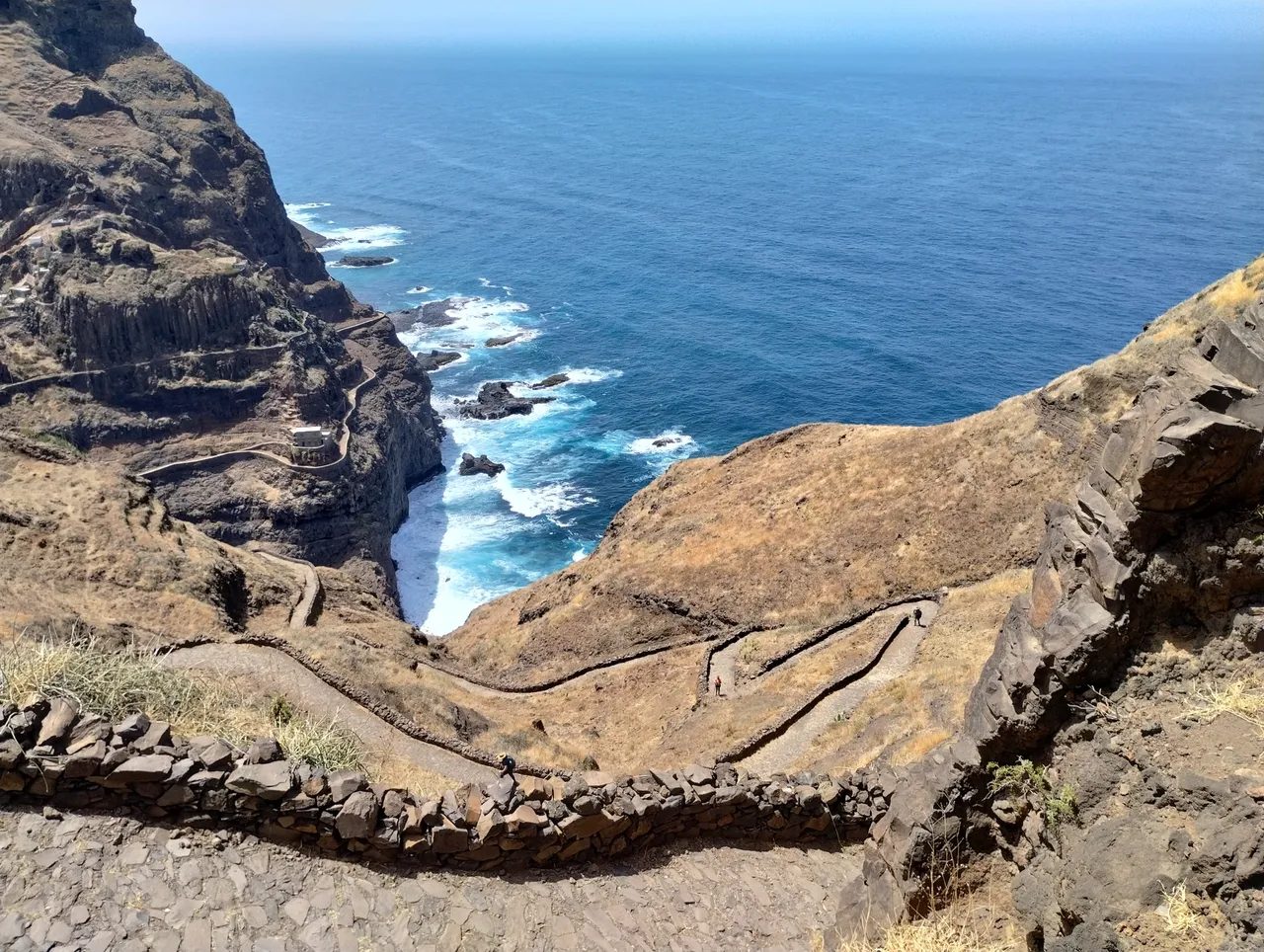 Stone-walled zigzag path descending a dry hillside towards a rocky sea inlet on the north coast of Santo Antão